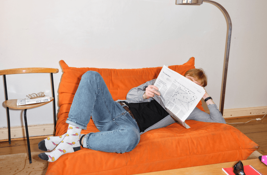 A person lying down on an orange couch, reading the newspaper.